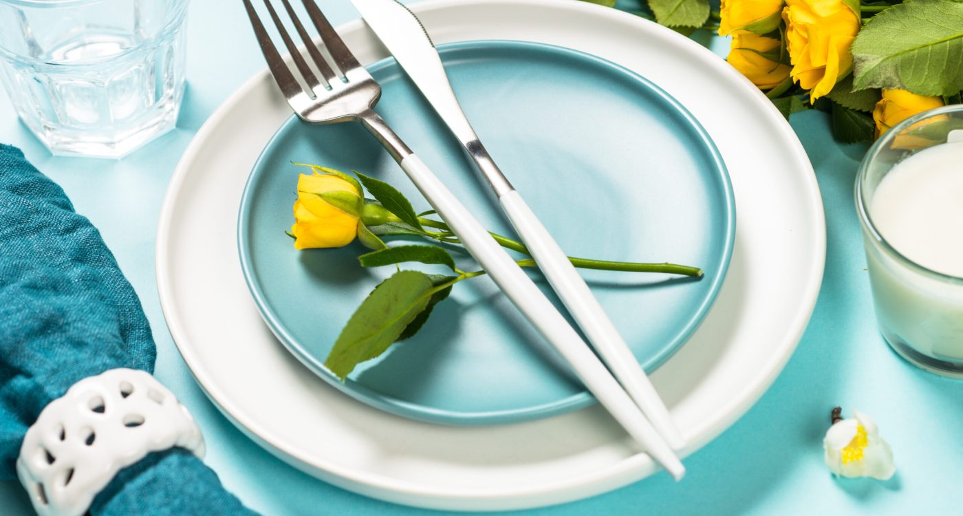 Spring table setting, served with rose flower, white plate and cutlery on blue background.
